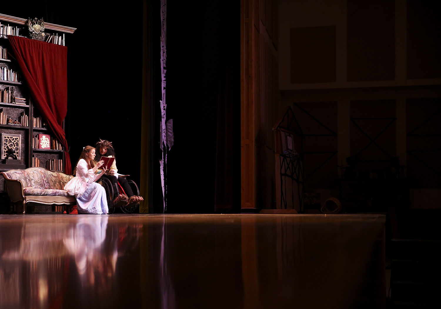 Kate Norman as Belle and Cooper Smith as the Beast sit together reading a book